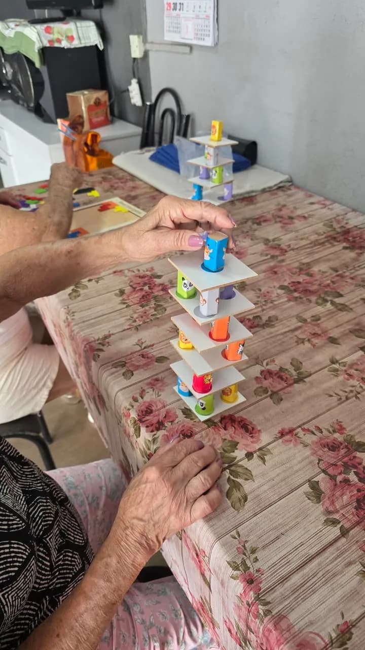 Mãos de idosa empilham blocos coloridos sobre toalha de mesa florida durante atendimento domiciliar.